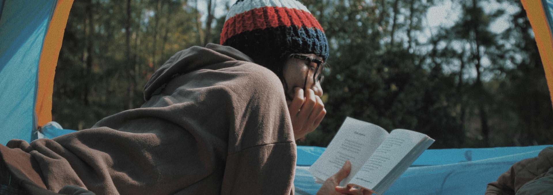 A lady in a wooly hat reading a book while out camping