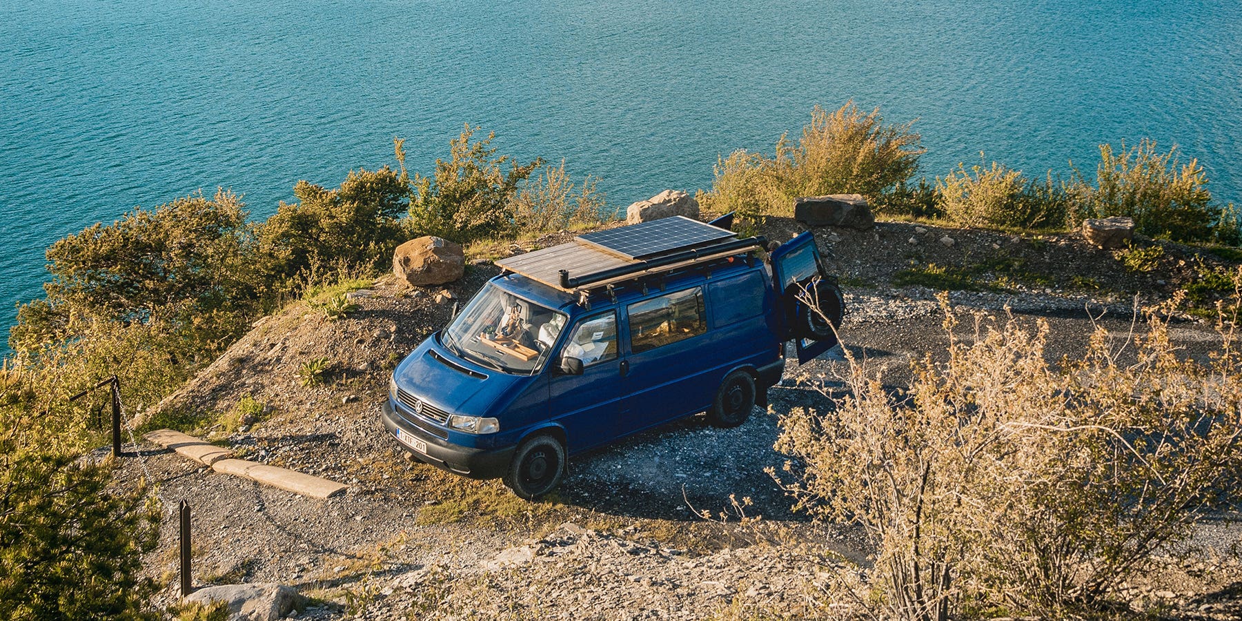 A blue VW T4 parked on a cliff by the sea, with a solar panel on the roof